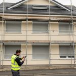 Façade d’une maison en rénovation avec échafaudage, volets roulants motorisés fermés et artisan contrôlant une télécommande.
