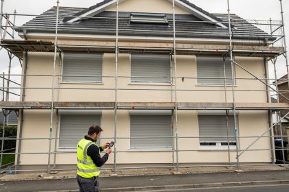 Façade d’une maison en rénovation avec échafaudage, volets roulants motorisés fermés et artisan contrôlant une télécommande.