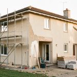 Maison individuelle en rénovation avec une façade partiellement restaurée, un échafaudage discret, des outils de chantier et une lumière naturelle.